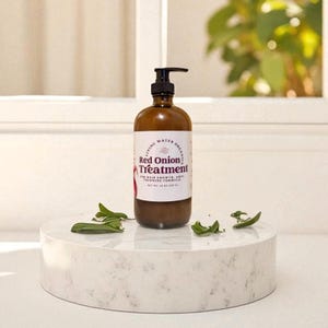 May include: A brown bottle of "Red Onion Treatment" for hair growth and anti-thinning formula. The bottle has a black pump dispenser and a white label with text. The bottle sits on a white, round, marble-like platform with green leaves.