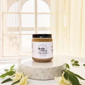 May include: A jar of "Scalp Exfoliating" scrub with a black lid and a white label. The label reads "Organic Cane Sugar & Jojoba Oil Scrub". The jar is on a white, speckled pedestal with greenery and flowers.