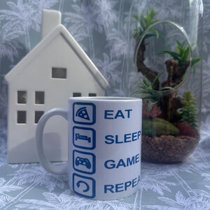 May include: White ceramic mug with the words "EAT SLEEP GAME REPEAT" in blue, and related icons. A white ceramic house and a glass terrarium with plants are in the background.