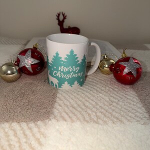 May include: White ceramic mug with a handle, featuring a teal forest design and the words "Merry Christmas." The mug is surrounded by red and gold Christmas baubles, including star-shaped and round decorations, and a small red reindeer figurine.