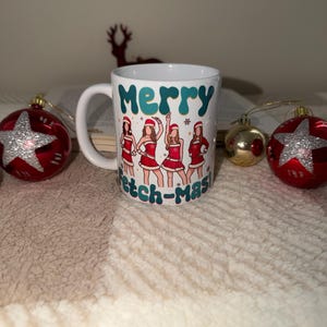 May include: White ceramic mug with the words "Merry Fetch-mas" and a graphic of four women in Santa hats and red dresses. The mug is surrounded by Christmas ornaments, including red star-shaped and gold round ornaments.