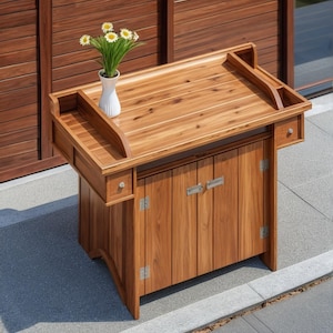 May include: A wooden outdoor bar cart with a top shelf and a cabinet with two doors. The cart has a white vase with white flowers on the top shelf.