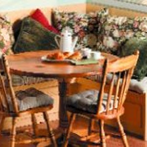 Può includere: Un angolo colazione con un tavolo rotondo in legno e quattro sedie. L'angolo ha posti a sedere integrati con cuscini e un tessuto a fantasia floreale. Il tavolo ha una caraffa, piatti e pasticcini.