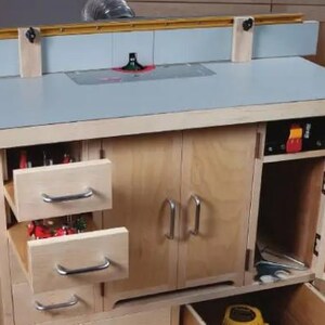May include: A wooden router table with a light blue top, two drawers, two doors, and a small cabinet with a door. The router table has a built-in fence and a router bit installed.