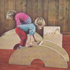 May include: Two children playing on wooden arch-shaped toys. One child is climbing on the arch, while the other is on top. The toys are light brown, and the floor is red. The background is a wooden wall.