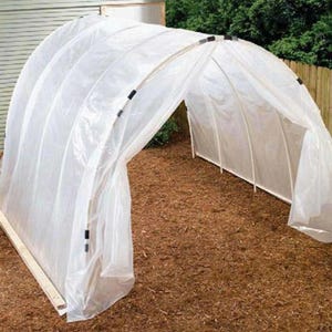 May include: A white plastic hoop house with a clear plastic cover. The arched structure is supported by white poles and secured with black clips. The hoop house is set on a bed of brown mulch, with a wooden frame at the base.