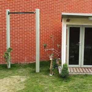 Puede incluir: Una barra de dominadas gris está montada entre dos postes grises claros frente a una pared de ladrillos rojos. Hierba verde y pequeñas plantas en primer plano. Un pequeño edificio con puertas de cristal a la derecha.