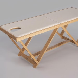 Könnte beinhalten: Helle Massageliege aus Holz mit cremefarbener, gepolsterter Oberfläche. Der Tisch hat eine Kopfstütze und ein zusammenklappbares X-Rahmen-Design. Der Tisch ist etwa 183 cm lang.