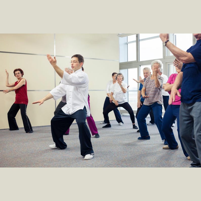 Puede incluir: Un grupo de personas practicando Tai Chi en interiores. Visten ropa holgada, incluyendo camisas blancas y pantalones negros. La habitaci&oacute;n tiene grandes ventanas y una combinaci&oacute;n de colores neutros.