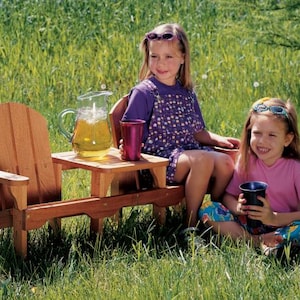 May include: Two children sit at a wooden picnic table with a pitcher of lemonade and two cups. The table and chairs are made of natural wood. The children are wearing casual summer clothing.