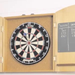 Puede incluir: Un armario de madera para dardos con una pizarra negra para marcar el marcador. La pizarra tiene los nombres "Scott" e "Ian" con las puntuaciones enumeradas debajo. El tablero de dardos es un tablero de dardos de marca "Puma" en blanco y negro con un centro rojo.