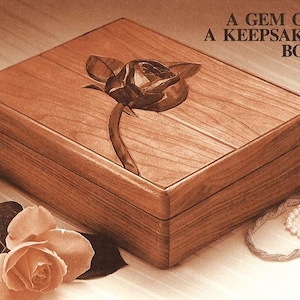 Puede incluir: Una caja de recuerdo de madera con un diseño de rosa tallada en la tapa. La caja está hecha de madera de cerezo y tiene un color marrón intenso. El texto "A GEM OF A KEEPSAKE BOX" está impreso en el fondo.