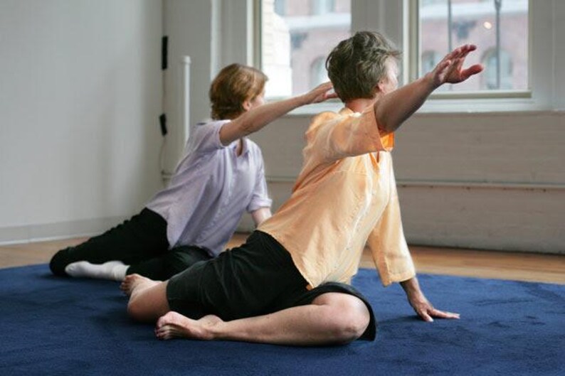 May include: Two people are practicing yoga on a blue mat. One person is wearing an orange shirt and black shorts, with their arm extended. The other person is wearing a light purple shirt and black trousers.