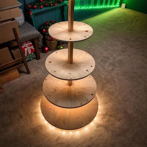 Op de afbeelding: Een houten displaystandaard met meerdere niveaus en vier ronde planken van verschillende afmetingen. De standaard is verlicht met kleine lampjes rond de basis. Het hout heeft een natuurlijke afwerking.