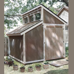 Puede incluir: Un cobertizo de madera marrón con un techo a dos aguas y una ventana abuhardillada. El cobertizo tiene puertas dobles y está rodeado de hierba verde y plantas en macetas.