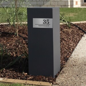 May include: A dark gray rectangular mailbox with a silver address plate. The plate displays the number "35" above the street name "Frederick St". The mailbox is set in a garden bed with mulch and plants.