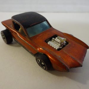 May include: An orange die-cast toy car with a black roof and black wheels. The car has a silver engine detail in the hood. The toy car is a vintage collectible.