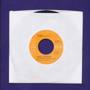 May include: A black vinyl record inside a white paper sleeve. The record features an orange center label with the text Dalida, Darla Dirladada, RCA, and Victor. The label includes production details, the orchestra name, and the SIAE stamp.