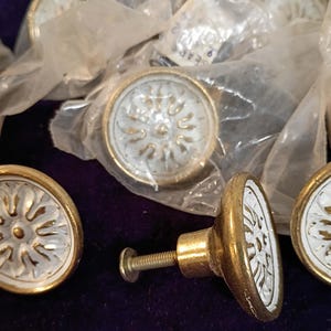 May include: Close-up of several vintage cabinet knobs. The knobs have a white center with a gold sunburst design and a gold rim. One knob is fully assembled with a screw. Some knobs are still in their original plastic packaging.