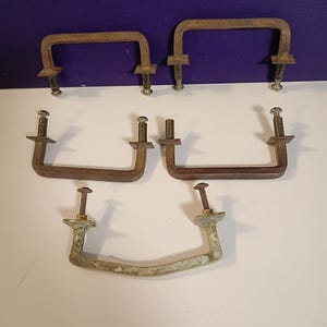 May include: A collection of vintage metal drawer pulls and handles. The pulls are in various shapes, including rectangular and curved designs, with visible signs of age and wear. The hardware has a rustic, antique appearance, with a mix of brown and green patina.