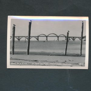 May include: Black and white vintage photograph of a bridge over water. Three wooden posts with birds on top are in the foreground. The bridge has multiple arches. Text at the bottom reads "TOUR OF A KIND - ALSEA BRIDGE, WALDPORT, OREGON."