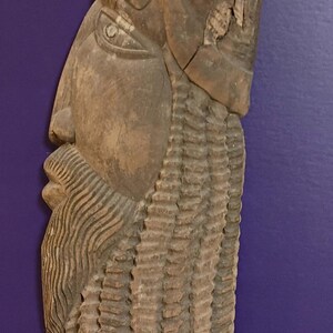 May include: A weathered, carved wooden sculpture of a bearded face in profile. The sculpture has a long, textured beard and a hat-like structure on top. The wood has a natural, aged appearance with visible grain and cracks.