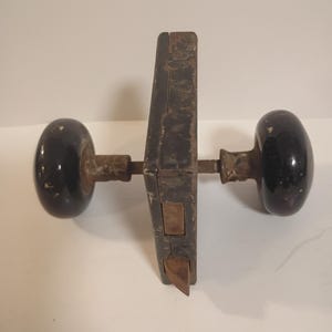 May include: An antique door lock set with black oval doorknobs and a dark metal lock mechanism. The lock shows signs of age and wear, with a rusty appearance. The doorknobs are glossy and reflect light.
