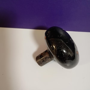 May include: A round, glossy black door knob with a smooth, reflective surface. The knob features a metallic, weathered base with visible signs of wear and small patches of rust, showing a cylindrical neck with a small hole for mounting hardware.