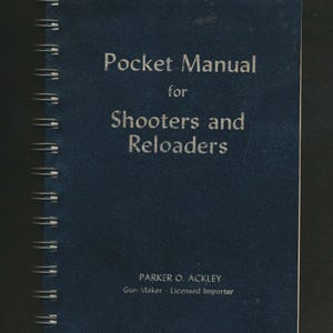 May include: A navy blue spiral-bound book titled "Pocket Manual for Shooters and Reloaders." The cover features silver lettering and the author's name, Parker O. Ackley, with the text "Gun Maker - Licensed Importer."