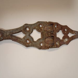May include: An antique, ornate metal hinge with a weathered, rusty appearance. The hinge features decorative cutouts and multiple holes for mounting. The metal has a mix of brown and gray tones, suggesting age and exposure to the elements. This hardware piece is likely for a door or gate.