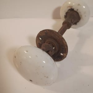 May include: Antique white porcelain door knob with a rusty metal base and stem. The oval-shaped knob has a smooth, glossy finish with small dark spots. The round base has three screw holes.