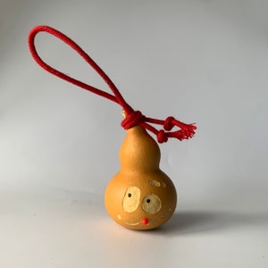 Könnte beinhalten: Eine kleine, braune Kalebasse mit einem bemalten Gesicht und einer roten Schnur, die um die Oberseite gebunden ist. Die Kalebasse hat zwei Augen, eine Nase und einen Mund.
