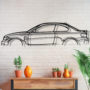 May include: Black metal wall art depicting a car silhouette, displayed against a white brick wall with potted plants and a wooden shelf below.