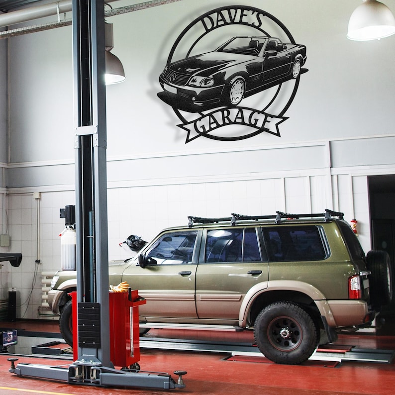 Op de afbeelding: Een zwart-wit metalen bord met een auto-silhouet en de tekst "Dave's Garage" in een cirkel. Het bord is gemonteerd op een witte muur in een garage.