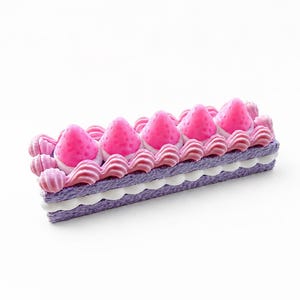 May include: A rectangular, miniature cake with layers of purple, white, and pink. The top is decorated with pink frosting, pink strawberry shapes, and decorative pink swirls. The cake sits on a white surface.