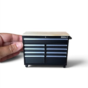 May include: A miniature black tool chest with a light wood-toned top surface and silver drawer handles. The front of the unit features the brand name KOBALT printed in white lettering on the upper right side, with multiple drawers arranged in a grid.