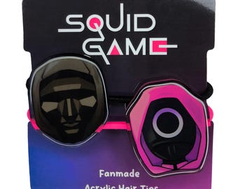 May include: Two acrylic hair ties featuring designs from Squid Game. One is a black mask, the other a pink and black guard. The hair ties are attached to a black card with the words "SQUID GAME" and "Fanmade Acrylic Hair Ties".