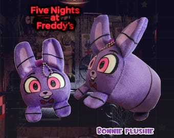 May include: Two purple Bonnie plushies from Five Nights at Freddy's. The plushies have large pink and white eyes, a red bow tie, and long ears. The text "Five Nights at Freddy's" and "Bonnie Plushie" are visible.