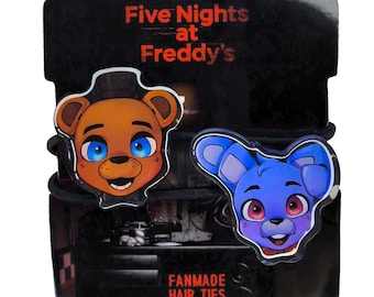 May include: Two black hair ties with a brown bear and a blue bunny character from the video game Five Nights at Freddy's. The packaging says "Fanmade Hair Ties".