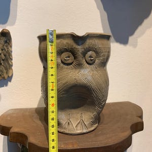 Op de afbeelding: Een handgemaakte, keramische uilvormige plantenbak, ongeveer 20 cm hoog. De plantenbak heeft een gedempte grijsbruine kleur met gesneden details die veren en gezichtskenmerken uitbeelden. Hij staat op een houten plank.