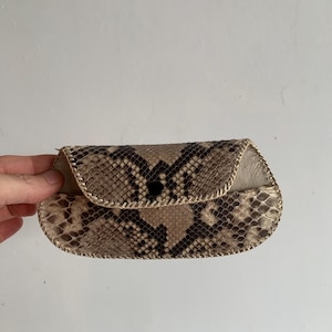 Könnte beinhalten: Kleine, handgefertigte Schlangenleder-Clutch mit gebogenem Überschlag und schwarzem Knopfverschluss. Die Clutch hat ein braun-beiges Schlangenmuster mit einer genähten Bordüre. Die Innenseite ist hellbeige.