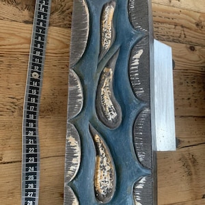 May include: A rectangular, carved wooden decorative piece with a blue and gold design. The piece is approximately 12 inches long and features a series of teardrop-shaped elements. The edges are dark gray, and the piece has a silver metal attachment.