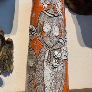 Op de afbeelding: Een hoge, oranje keramische vaas met een textuur. De vaas heeft een reliëfontwerp van een vrouw en een kind, met witte en grijze details. De vaas is een decoratief item.