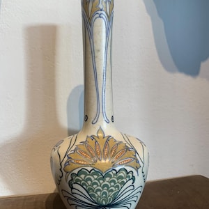 Pode incluir: Um vaso de cerâmica branco sujo com um pescoço fino e uma base bulbosa. O vaso é adornado com padrões florais em tons de amarelo, verde e azul. O design apresenta uma grande flor na base e motivos florais menores ao longo do pescoço.