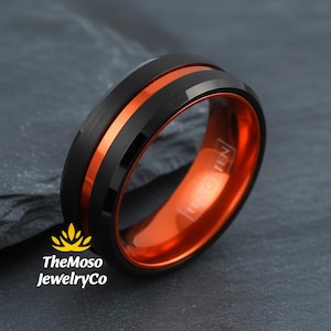 May include: Black and orange tungsten ring. The ring features a black exterior with a central orange stripe and an orange interior. The ring is a close-up shot on a dark gray surface. TheMoso JewelryCo logo is in the lower left corner.