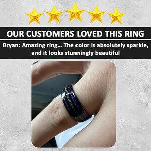 May include: A black ring with a multi-colored inlay. The inlay has shades of purple, blue, and silver. The ring is worn on a person's finger.