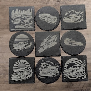 May include: Set of ten black slate coasters with white designs of cars and cityscapes. The coasters feature various designs, including a cityscape, a sunset, and a car with the word "JDM" on it.