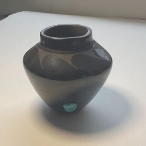 May include: A small, dark gray ceramic vase with a rounded body and a narrow opening. The vase has a turquoise stone accent on the side. The vase is likely handmade and has a smooth, glossy finish.