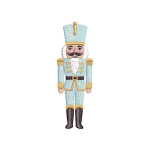 May include: Embroidered light blue nutcracker soldier with gold accents, a black belt, and black boots. The nutcracker has a white beard and mustache, and a gold-colored hat with a gold finial. A festive holiday decoration.