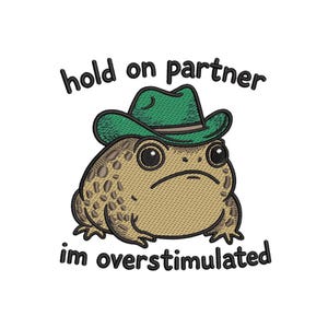 Puede incluir: Diseño bordado de una rana de dibujos animados con un sombrero de vaquero verde. La rana tiene una expresión de mal humor y es de color beige con manchas marrones. El texto de arriba dice "hold on partner" y abajo "im overstimulated".